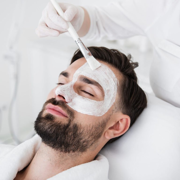 Cosmetologist using brush while putting white clay mask on male face